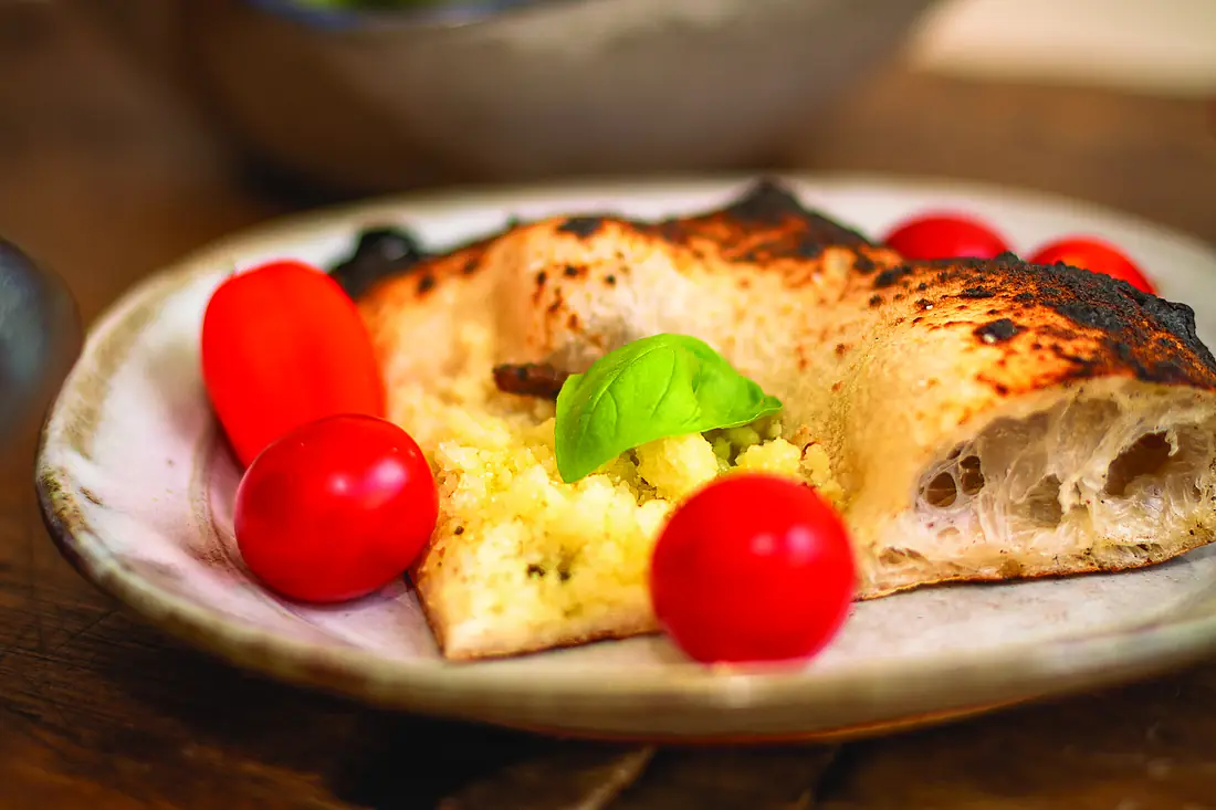 Porción de pizza napolitana cortada, mostrando el alveolado abierto del borde, junto a unos tomates.