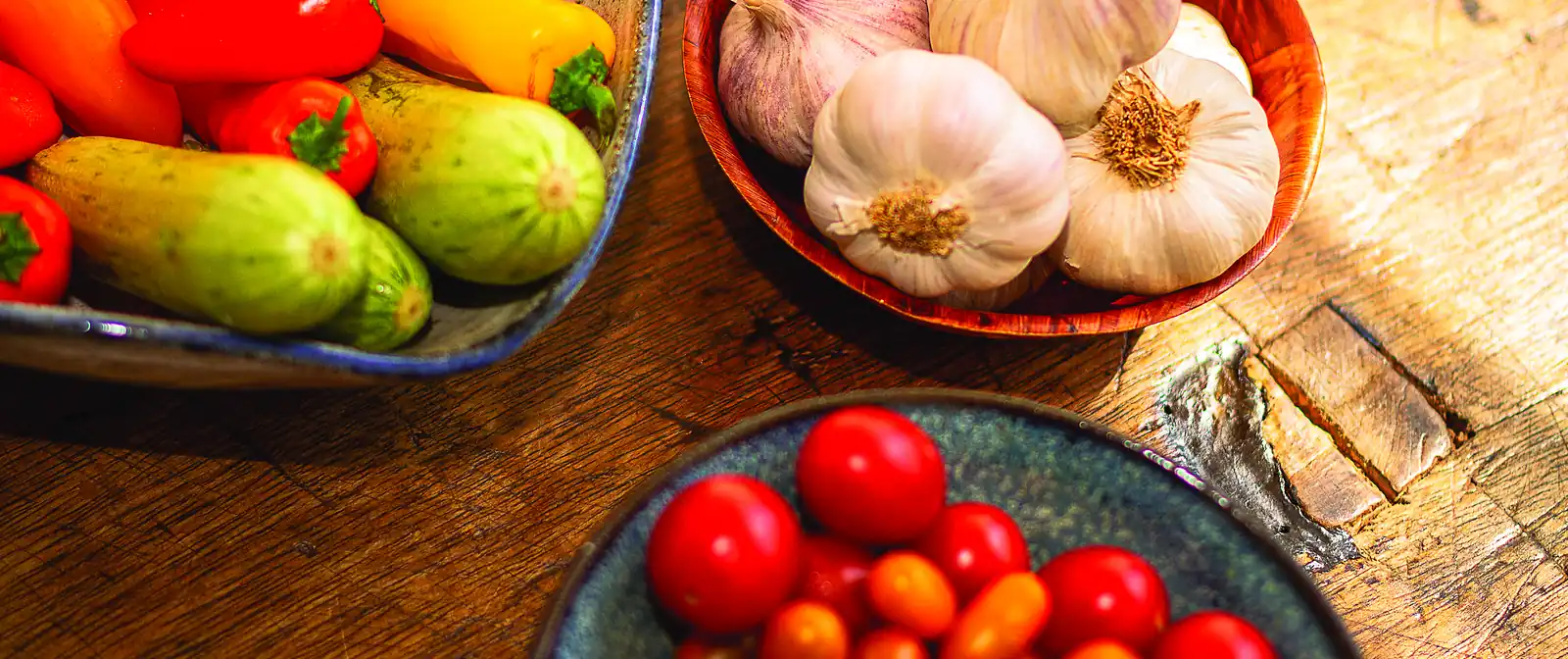Pimientos, pepinos, cabezas de ajo y tomates cherry en cuencos sobre una encimera de madera.
