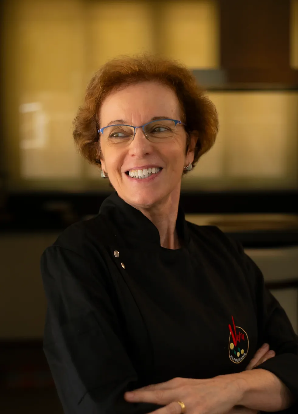 Vera Lucia Cegal, con chaquetilla negra y gafas, sonriendo de brazos cruzados en la cocina.